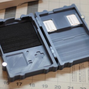 May include: A gray, hinged storage case, open to reveal two compartments. One compartment has a black foam insert, and the other holds two white cards labeled "SAMSUNG". The case has a retro game controller design.