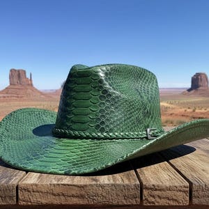 Może przedstawiać: Zielony kapelusz kowbojski ze wzorem skóry węża leży na drewnianej powierzchni. Kapelusz ma plecioną opaskę i srebrną klamrę. Tło przedstawia pustynny krajobraz z mesami.