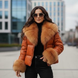 Puede incluir: Chaqueta acolchada marrón claro con cuello y puños de piel. La chaqueta se lleva sobre un cuello alto negro y vaqueros negros. El fondo presenta un paisaje urbano borroso.