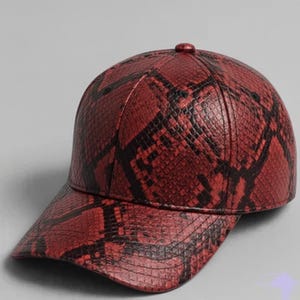 Może przedstawiać: Czerwono-czapka bejsbolówka z czarnym wzorem wężowej skóry. Czapka ma zakrzywiony daszek i guzik na górze. Wykonana z teksturowanego materiału, prawdopodobnie ze skóry ekologicznej.