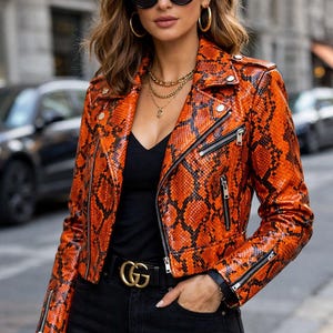 Pode incluir: Casaco laranja e preto com padrão de pele de cobra, zíperes prateados e cinto preto com fivela dourada. O casaco tem um estilo motociclista clássico com gola entalhada e silhueta ajustada. A pessoa usa óculos de sol pretos e joias douradas.