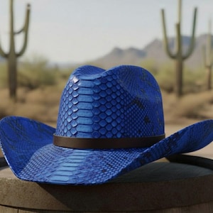 Może przedstawiać: Niebieski kapelusz kowbojski z wzorem skóry węża i brązowym skórzanym paskiem. Kapelusz leży na drewnianym beczce, z krajobrazem pustynnym w tle.