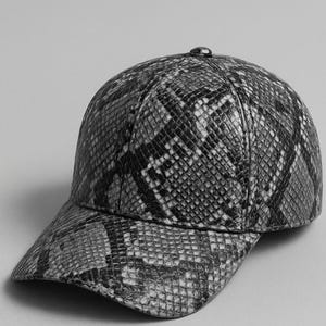Könnte beinhalten: Baseballkappe mit Schlangenmuster in Grau- und Schwarztönen. Die Kappe hat einen gebogenen Schirm und einen kleinen Knopf oben. Der Hut besteht aus einem strukturierten Material.