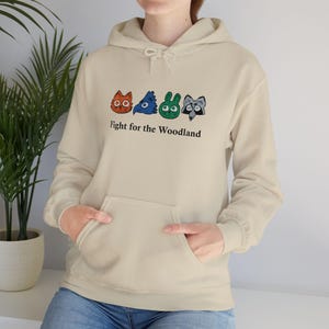 Puede incluir: Sudadera con capucha beige con bolsillo delantero y cordón. Presenta un gráfico colorido de cuatro caras de animales del bosque sobre el texto "Fight for the Woodland". Las caras de los animales son naranja, azul, verde y gris.