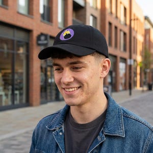 May include: Black baseball cap with a curved brim. The cap features a circular patch with a yellow and black design on a purple background. The person in the image is wearing a denim jacket and a gray t-shirt.