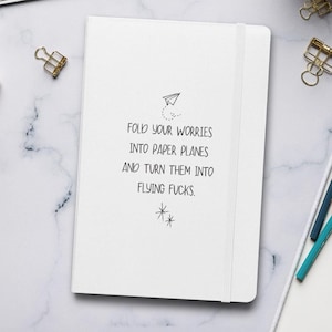 May include: White notebook with the text "Fold your worries into paper planes and turn them into flying f*cks." The notebook has a paper airplane illustration and a matching elastic band closure. Gold paper clips are visible.
