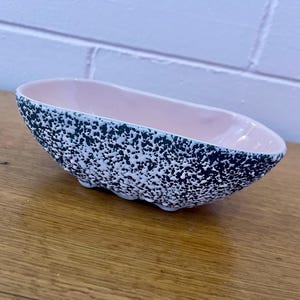 May include: A pink and black speckled ceramic bowl with a rounded oval shape. The bowl has a textured surface and a smooth, glossy finish.