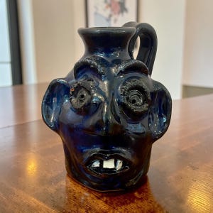 May include: A dark blue ceramic jug with a face sculpted on the front. The jug has a handle and a spout. The face has large, textured eyes, a wide mouth with two white teeth, and large ears. The jug is sitting on a wooden surface.