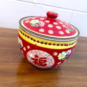 May include: A red ceramic jar with a lid, featuring a floral design and the Chinese character for "good fortune." The jar has a yellow band with a diamond pattern and a small red knob on top. The jar is on a wooden surface.
