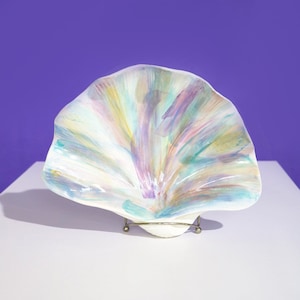 May include: A large, white ceramic seashell with a iridescent finish. The shell is painted with shades of blue, green, purple, and yellow. It is displayed on a metal stand.