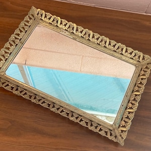 May include: A rectangular vanity mirror with an ornate, gold-colored frame. The frame features a detailed, repeating pattern around the edges. The mirror reflects a light blue and pink hue. The mirror is placed on a dark brown wooden surface.