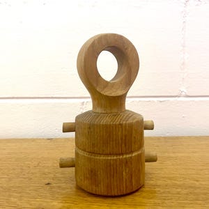 May include: A light brown wooden object with a circular handle and two small wooden pegs on each side. The object is sitting on a wooden surface. The background is a white wall.