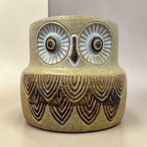May include: A ceramic owl-shaped container in shades of tan and brown. The owl has large, detailed eyes with white and brown accents. The body features a textured pattern of downward-pointing, stylized feathers. A decorative piece.