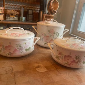 May include: A set of three white enamel pots with matching lids, each featuring a delicate floral pattern in shades of pink, green, and blue. The pots have two side handles and a curved lid handle, showcasing a smooth, glossy finish throughout.