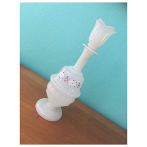 May include: A white glass vase with a tulip-shaped top. The vase has a bulbous base and a slender neck, with a decorative band of red and white floral accents. The vase is set against a blue background.