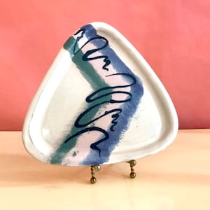 May include: A triangular ceramic serving tray with a white background and abstract blue, green, and pink painted design. The tray is on a gold metal stand.