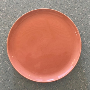 May include: A round, coral-colored plate. The plate has a smooth surface and a slightly raised edge. The plate is centered on a light blue surface with a repeating pattern of white and black curved lines. This plate is suitable for serving food.