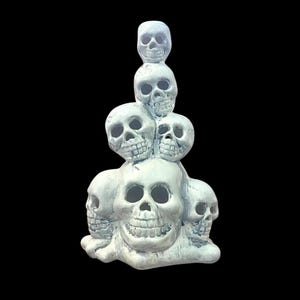 May include: A stacked sculpture of skulls in shades of gray. The sculpture features a base of multiple skulls, with additional skulls stacked on top, creating a towering effect. The skulls have detailed features and are set against a black background.