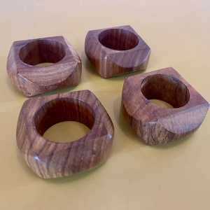 May include: Four wooden napkin rings in a square shape with rounded edges. The rings are made of dark brown wood with a natural grain pattern.
