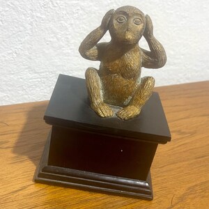 May include: A gold monkey figurine with its hands over its ears, sitting on a black wooden base. The monkey is part of a set representing the "See No Evil, Hear No Evil, Speak No Evil" proverb.
