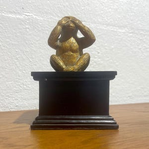 May include: A gold monkey figurine with its hands over its eyes, sitting on a black pedestal. The monkey is part of the "See No Evil" series.