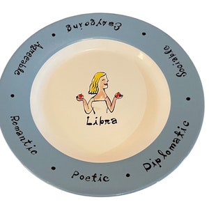 May include: A light blue ceramic plate with a white inner rim. The plate features a cartoon illustration of a woman with blonde hair holding two red apples. The text "Libra" is written below the illustration. The rim of the plate has the words "Sociable", "Charming", "Accessible", "Romantic", "Poetic", and "Diplomatic" written around it.