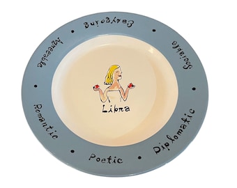 May include: A light blue ceramic plate with a white inner rim. The plate features a cartoon illustration of a woman with blonde hair holding two red apples. The text "Libra" is written below the illustration. The rim of the plate has the words "Sociable", "Charming", "Accessible", "Romantic", "Poetic", and "Diplomatic" written around it.