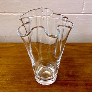 May include: A clear glass vase featuring a wide, undulating, ruffled rim that creates a series of soft, organic curves. The body of the vase tapers downward from the flared top to a solid, circular base, showcasing a transparent, smooth finish.