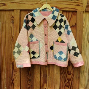 Pu&ograve; includere: Giacca trapuntata rosa con un design patchwork a rombi neri, blu e bianchi. La giacca ha un colletto, tasche e bottoni. &Egrave; appesa a una gruccia di legno.