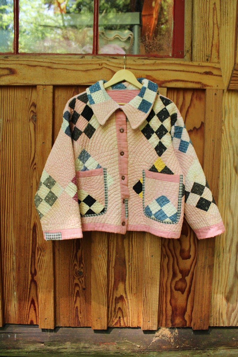 Pu&ograve; includere: Giacca trapuntata rosa con un design patchwork a rombi neri, blu e bianchi. La giacca ha un colletto, tasche e bottoni. &Egrave; appesa a una gruccia di legno.