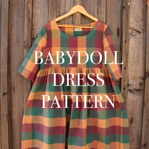 Puede incluir: Un patrón de vestido babydoll a cuadros en tonos verdes, naranjas y burdeos cuelga de una percha de madera. El vestido presenta un escote redondo y mangas cortas. Las palabras "BABYDOLL DRESS PATTERN" están impresas en la parte delantera del vestido.