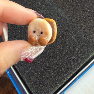 Könnte beinhalten: Eine Miniatur-Sandwich-Figur mit einem Lächeln. Das Sandwich hat eine hellbraune Brotaußenseite, eine braune Füllung und braune Arme und Beine. Die Figur wird zwischen Fingern mit einem verzierten Nagel gehalten.