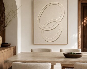 Könnte beinhalten: Ein rechteckiges Wandbild mit einer strukturierten beigen Leinwand, auf der zwei große, sich überschneidende ovale Formen in einem erhabenen, einfarbigen Relief dargestellt sind. Das Kunstwerk ist in dünnes helles Holz gefasst.