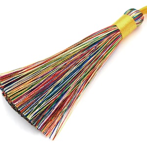 May include: A vibrant tassel featuring a yellow band and loop. The tassel is composed of numerous thin, multi-coloured threads, including red, blue, green, and yellow. The threads are tightly bound at the top and spread out towards the bottom.