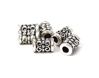 Pode incluir: Uma coleção de contas de metal prateado. Cada conta cilíndrica apresenta um design ornamental e simétrico com um motivo floral central e detalhes texturizados. As contas estão espalhadas sobre um fundo branco.
