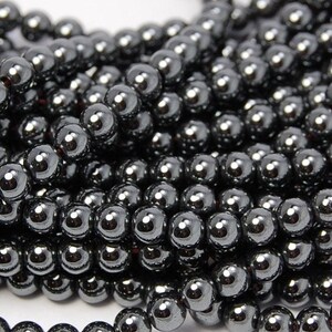 Peut inclure: Un brin de perles d'hématite noire, chaque perle est ronde et lisse. Les perles sont disposées en gros plan, montrant la finition brillante et métallique.