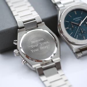 Op de afbeelding: Twee roestvrijstalen horloges. Eén horloge heeft een teal-kleurige wijzerplaat met de tekst "architect London". Het andere horloge heeft de tekst "Jackson Your Time, Your Story" gegraveerd op de achterkant.