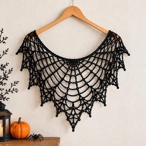 May include: A black crochet shawl featuring an intricate spider web pattern with a scalloped edge. The garment is displayed on a wooden hanger, showcasing the detailed knotted yarn construction and the pointed, symmetrical design of the web.