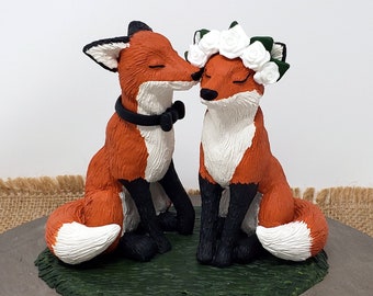 Può includere: Una volpe rossa con un papillon nero bacia una volpe rossa con una corona di fiori bianchi. Le volpi sono sedute su una base verde. La base è su un supporto per torte in legno. Il testo "My Custom Cake Topper" è sul supporto per torte.