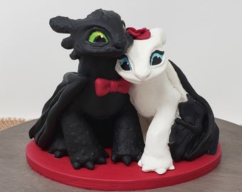 Può includere: Un topper per torta a forma di drago bianco e nero con un papillon rosso e un fiore rosso. I draghi sono in una posa romantica, con la testa del drago nero che riposa sulla spalla del drago bianco. Il topper per torta è su una base rossa.