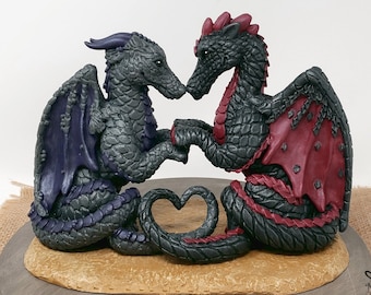Può includere: Un topper per torta a forma di drago nero e rosso con ali viola e rosse. I draghi si tengono per mano e le loro code formano un cuore.