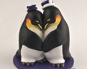 Può includere: Un topper per torta con due pinguini su una base circolare viola. Un pinguino indossa un cilindro bianco con una fascia viola, l'altro ha un fiore viola con un velo bianco. Il testo My Custom Cake Topper è visibile.