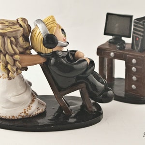 May include: A wedding cake topper featuring a bride pulling a groom sitting in a chair in front of a computer desk. The groom is wearing a black suit and headphones. The bride is wearing a white dress. The computer desk is brown with a black computer monitor and a black tower.
