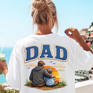 May include: A white t-shirt featuring a large blue embroidered-style text that reads DAD. Below the text is a graphic of an adult and a child sitting together, facing a large orange sun, with clouds, birds, and green grass in the background.
