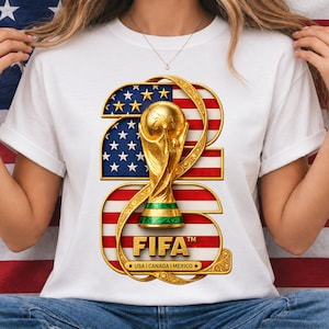May include: A white t-shirt featuring a large graphic of the number 2026 in gold, filled with the United States flag pattern. A gold FIFA World Cup trophy sits in the center. The text FIFA and USA I CANADA I MEXICO are printed in gold.