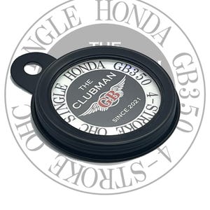 May include: A black metal motorcycle part with the text "SINGLE HONDA THE CLUBMAN GB350 4-STROKE" in a circular design. The design includes a winged emblem and the text "SINCE 2021".