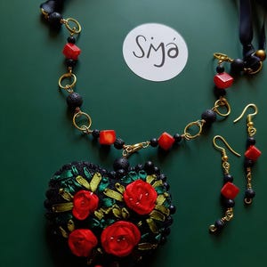 Puede incluir: Un conjunto de joyas hecho a mano con un colgante en forma de corazón con bordados de rosas rojas, cuentas de lava negras y detalles de cubos rojos. El conjunto incluye un collar y pendientes a juego, sobre un fondo verde oscuro.