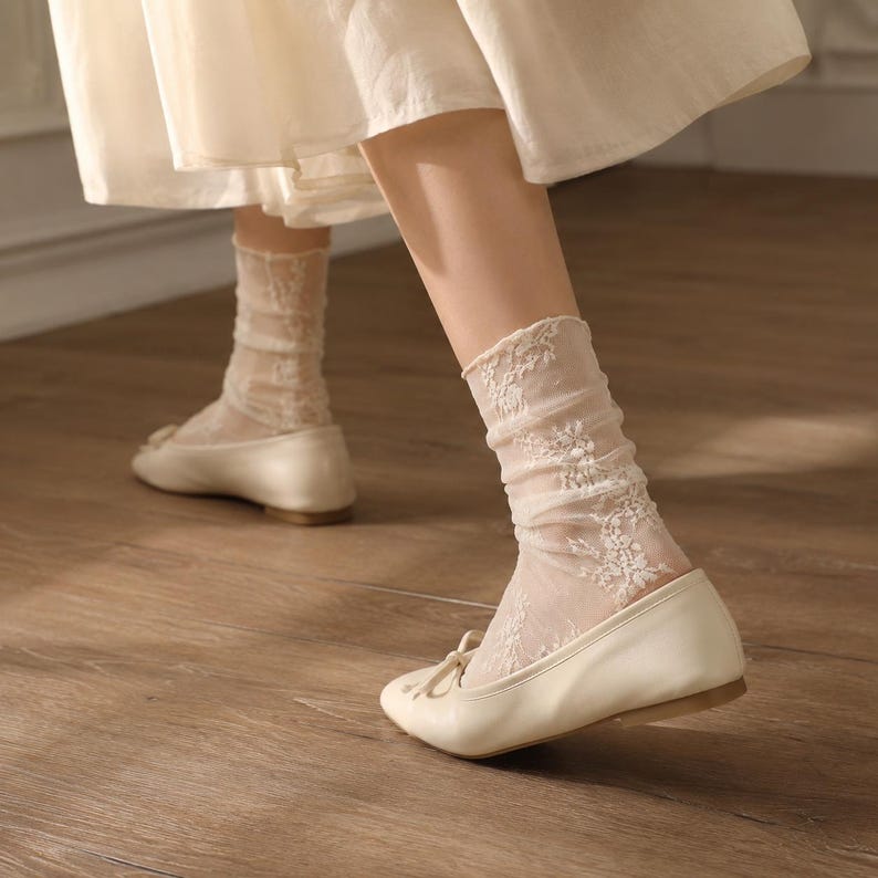 K&ouml;nnte beinhalten: Ein Paar cremefarbene Ballerinas mit einer kleinen Schleife vorne, getragen mit transparenten, wadenhohen Socken mit einem zarten wei&szlig;en Blumen-Spitzenmuster. Die Socken sind am Kn&ouml;chel leicht gerafft und haben einen gewellten Rand.