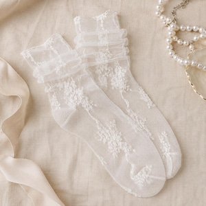 K&ouml;nnte beinhalten: Ein Paar halbtransparente, wei&szlig;e S&ouml;ckchen aus feinem Netzstoff. Die Socken sind mit einem komplexen floralen Spitzenmuster versehen und verf&uuml;gen &uuml;ber einen ger&uuml;schten, gerafften Abschluss am oberen Rand, was f&uuml;r eine strukturierte Optik sorgt.