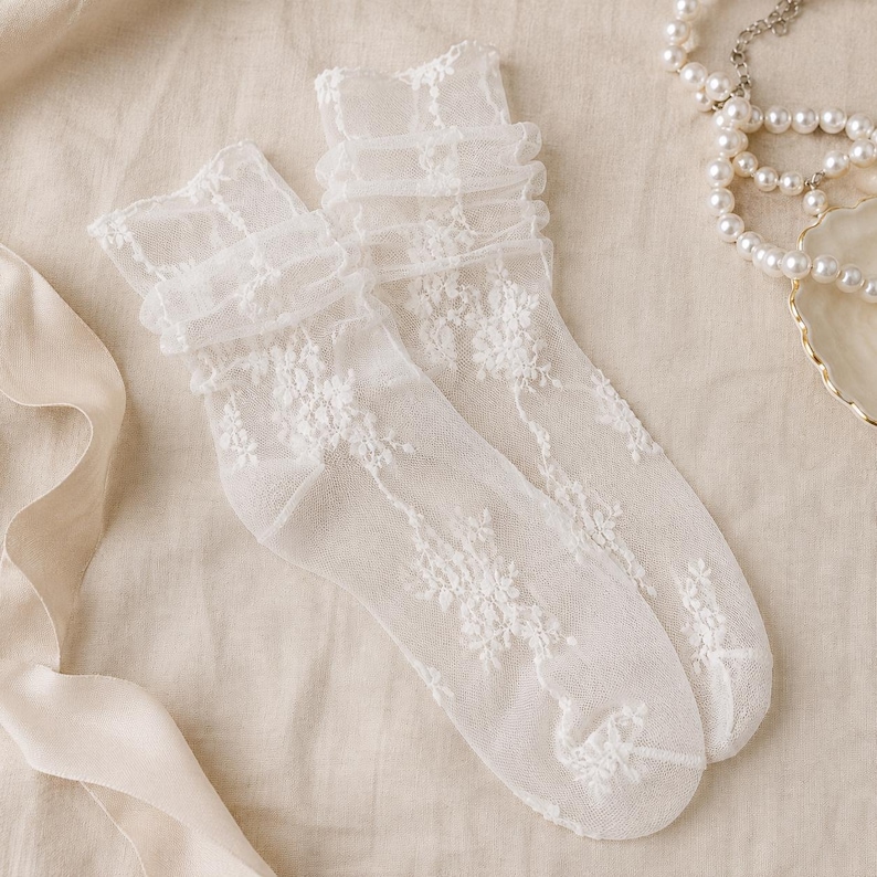K&ouml;nnte beinhalten: Ein Paar halbtransparente, wei&szlig;e S&ouml;ckchen aus feinem Netzstoff. Die Socken sind mit einem komplexen floralen Spitzenmuster versehen und verf&uuml;gen &uuml;ber einen ger&uuml;schten, gerafften Abschluss am oberen Rand, was f&uuml;r eine strukturierte Optik sorgt.